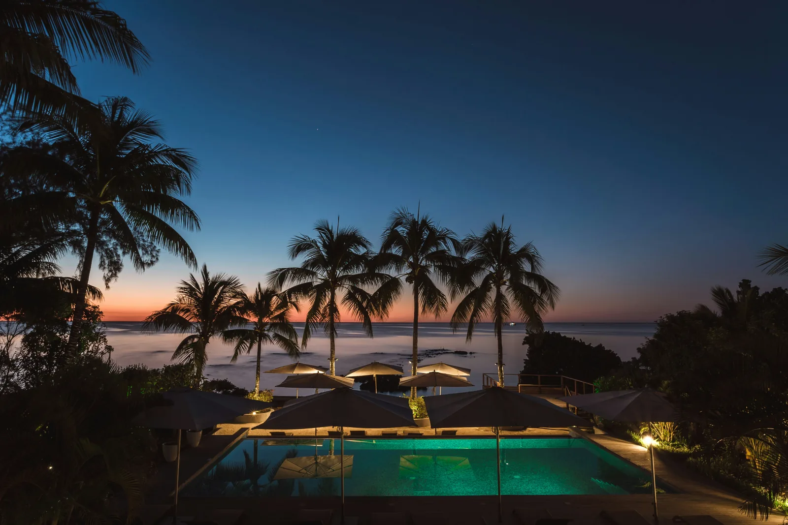 Praia de resort ao entardecer com coqueiros e guarda-sóis iluminados