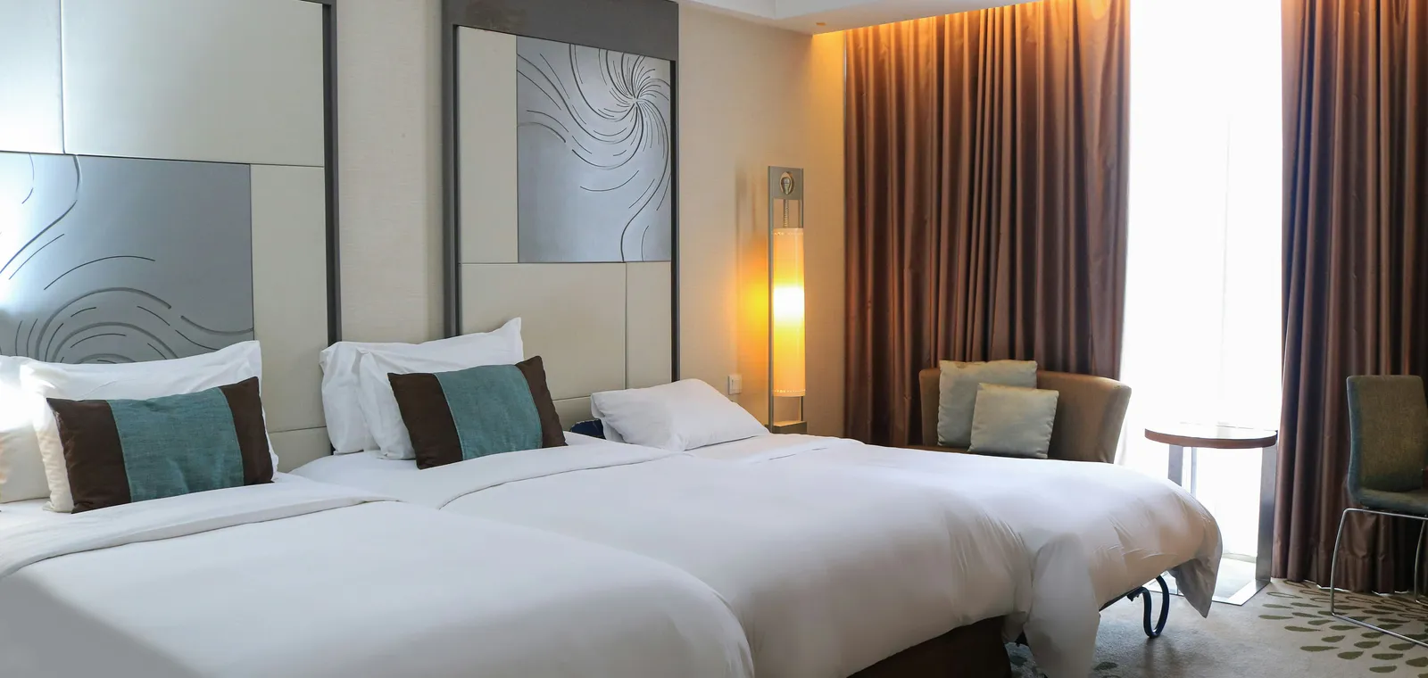 Quarto de hotel com camas de solteiro, cabeceiras em painéis decorativos e luz natural entrando pela janela